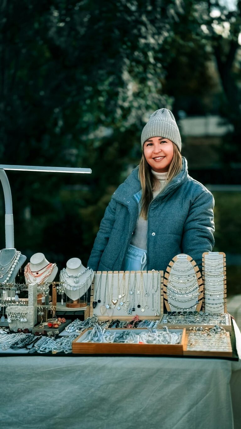 Smiling woman at outdoor jewelry stall in winter attire offering various jewelry.